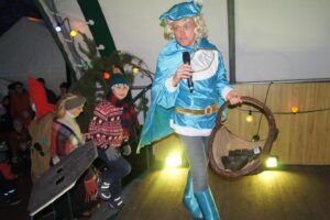 Eine Frau in einem blauen Kostüm hält ein Mikrofon und einen Wreath. Sie scheint Teil einer Veranstaltung zu sein.